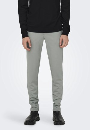 Mand iført smalle lysegrå bukser, sort langærmet top og sorte læderstøvler, stående med hænderne afslappet ned langs siderne.