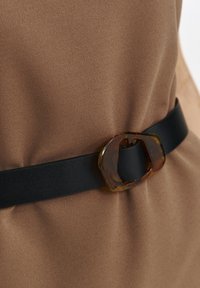 Czarny pasek z prostokątną klamrą z żółwia, noszony na brązowej fakturowanej tkaninie. Klamra ma przezroczysty, ziemisty wzór.