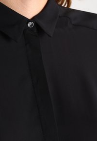 Camicia nera con colletto caratterizzata da un tessuto liscio e opaco. La parte anteriore presenta una chiusura a bottoni discreta e una linea elegante, creando un aspetto su misura.