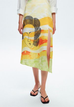 Żółta i pomarańczowa spódnica midi tie-dye z czarnym abstrakcyjnym nadrukiem twarzy i bocznym rozcięciem, noszona z czarnymi sandałami i białą koszulą.