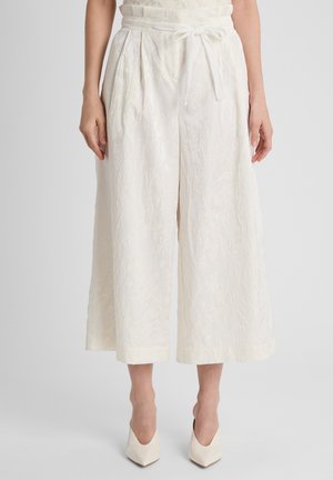 Vrouw draagt witte getextureerde cropped broek met hoge taille en wijde pijpen, met een bindceintuur en witte pumps met spits toelopende neus, staand op een grijze vloer.