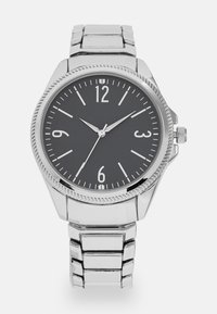 Montre analogique en argent avec un cadran noir, de grands chiffres blancs et un bracelet en acier inoxydable poli. Présente une lunette texturée et une couronne.