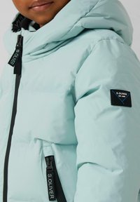 Šviesiai mėlynas pamuštas paltas su kapišonu, turintis juodą užtrauktuką ir logotipą ant rankovės. Lygus paviršius ir naudingos kišenės.