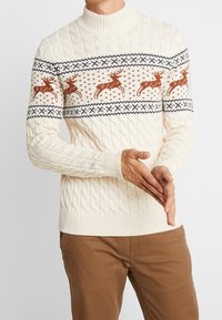 Krämmfärgad kabelstickad tröja med hög krage, försedd med bruna renmönster och blå detaljer över bröstet. Ribbstickade ärmslut och nederkant.