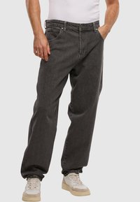 Man in losse donkergrijze jeans, witte sneakers en een witte mouwloze shirt, staand met handen gedeeltelijk in de zakken.