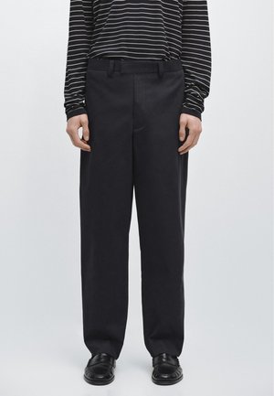 Persona che indossa pantaloni neri a gamba larga, mocassini neri e una camicia nera a maniche lunghe con righe orizzontali bianche, in piedi su uno sfondo semplice.