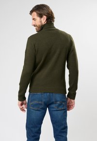 Pulover turtleneck de culoare verde închis, cu maneci lungi, material texturat și talie ajustată, asortat cu blugi albaștri care evidențiază detalii ale buzunarelor.