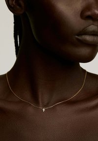 Collar de cadena de oro con un pequeño colgante de cruz, reposando sobre un tono de piel oscura. Diseño simple, textura fina y apariencia minimalista.