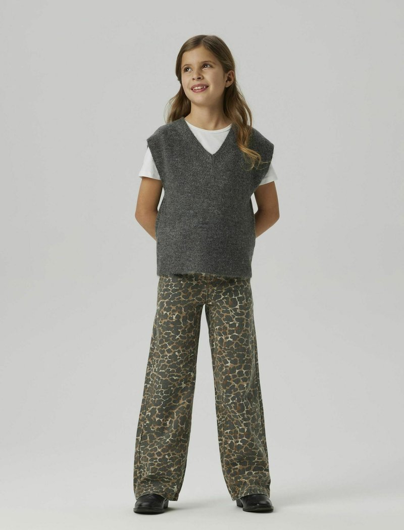 Un gilet en tricot gris avec un col en V sur un t-shirt blanc, associé à un pantalon large avec un imprimé léopard marron et vert. Chaussures noires.