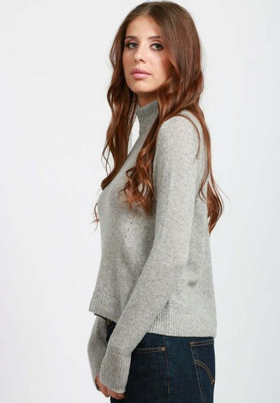 Donna con lunghi capelli castani ondulati che indossa un pullover a collo alto grigio chiaro e jeans blu scuro, in piedi di lato su uno sfondo semplice.
