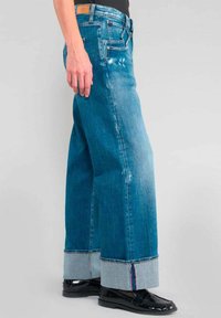 Jeans de mezclilla azul de pierna ancha con dobladillos deshilachados, que presentan puños de mezclilla clara en contraste y un ligero desgaste. Se ve el bolsillo delantero.