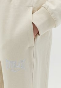 Beige Sport-Jogginghosen aus weichem Baumwollmix mit einer Seitentasche und einem weißen gestickten "Everlast"-Logo am linken Oberschenkel.