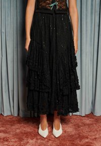 Čierna čipkovaná sukňa s vrstvenými volánikmi, zdobená flitrami. Kombinovaná s bielymi špicatými topánkami. V pozadí sa nachádza modré záclonové pozadie.