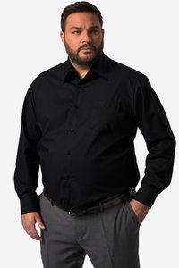 Mand med skæg iført en sort skjorte med knapper og grå bukser, stående med den ene hånd i lommen mod en ensfarvet baggrund.
