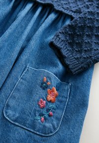 Robe en denim avec un haut en tricot bleu marine et une jupe bleue froncée. La poche présente une broderie florale colorée dans des teintes orange et bleue.