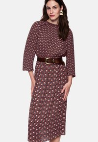 Bordeauxrotes Kleid mit geometrischen Mustern in Rot und Weiß; lange Ärmel, geraffter Halsausschnitt, tailliert mit braunem Gürtel und goldener Schnalle.