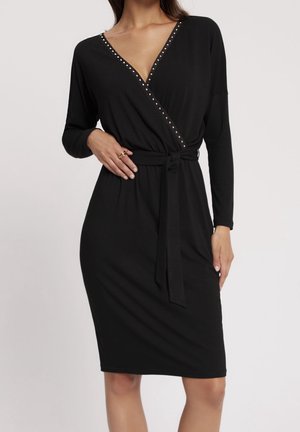 Czarna, długorękawowa sukienka kopertowa z dekoltem w kształcie litery V zdobionym ćwiekami oraz przewiązanym paskiem w talii, noszona przez modelkę z pierścieniami na lewej ręce.