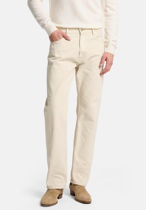 Beige Baumwolljeans mit geradem Schnitt, Fünf-Taschen-Design und sichtbaren Nähten. Getragen mit hellbraunen Wildlederstiefeln und einem cremefarbenen Pullover.