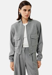 Szara bomberka wykonana z teksturowanej tkaniny, z zamkiem z przodu, dwiema przednimi kieszeniami z klapą oraz ściągaczami przy mankietach i u dołu.