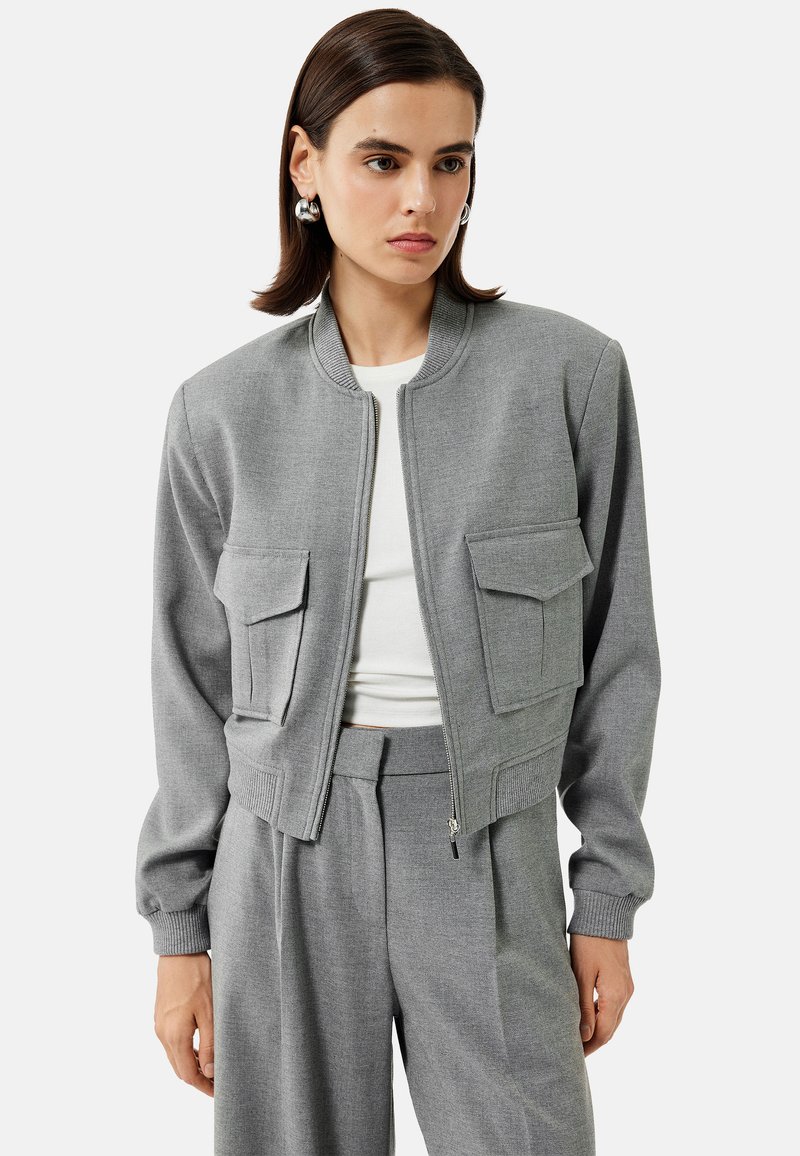 Szara bomberka wykonana z teksturowanej tkaniny, z zamkiem z przodu, dwiema przednimi kieszeniami z klapą oraz ściągaczami przy mankietach i u dołu.