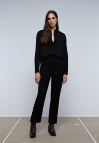 Camisa negra de botones con un sutil patrón texturizado, combinada con pantalones negros ajustados. El modelo lleva botas de tobillo, en un fondo neutro.