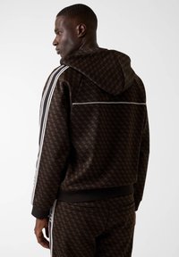 Brązowa bluza z kapturem w wzory, z białymi akcentami, wyposażona w kaptur ze sznurkiem i paskowane rękawy, wykonana z miękkiego materiału tekstylnego.