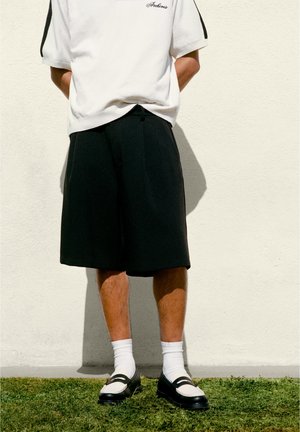 Person trägt ein weißes Kurzarmhemd, schwarze knielange Shorts, weiße Sportsocken und schwarz-weiße Loafers und steht auf Gras neben einer weißen Wand.