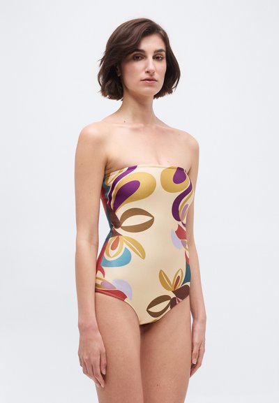 Γυναίκα με κοντά καστανά μαλλιά που φοράει ένα μπεζ μαγιό χωρίς τιράντες με αφηρημένα πολύχρωμα λουλουδένια σχέδια, στέκεται μπροστά σε ένα απλό λευκό φόντο.