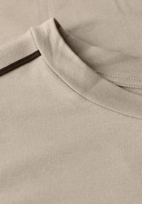 Nahaufnahme eines beige Farbtuchs, das den gerippten Ausschnitt und die Schulternaht mit einem strukturierten Besatzdetail zeigt.