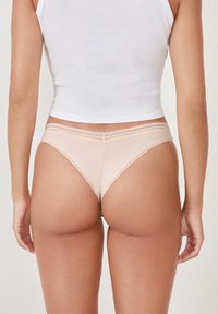 String beige avec un design texturé et côtelé, doté d'une dentelle à la taille. Le vêtement a une coupe basse.