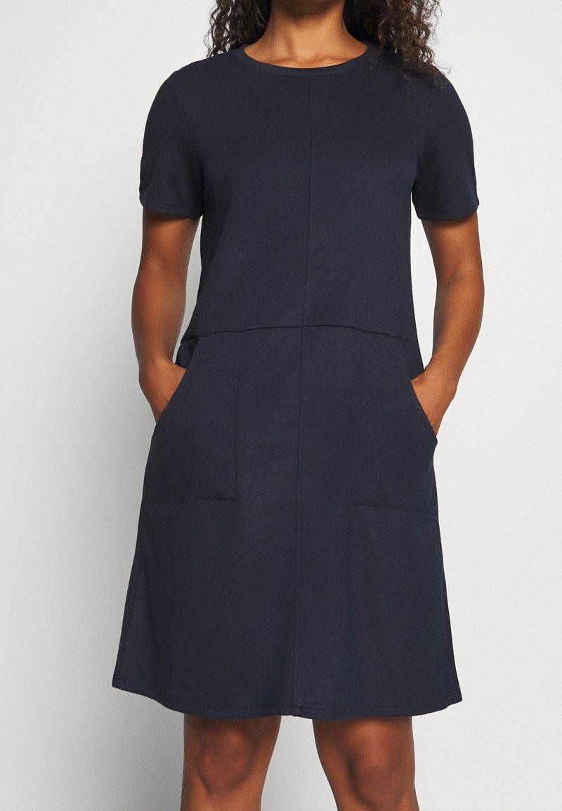 Femme portant une robe bleu marine à manches courtes, longueur genou, avec des poches sur le devant, debout les mains dans les poches sur un fond uni.