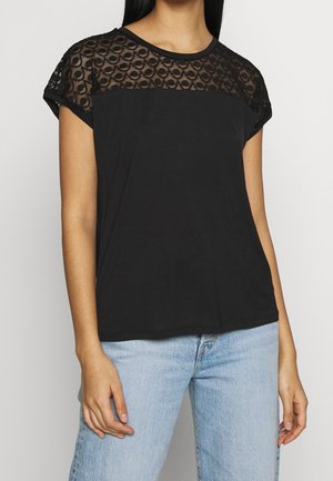 Kobieta ubrana w czarną bluzkę z krótkim rękawem z okrągłym wzorem koronki na górnej części klatki piersiowej i ramionach, w połączeniu z jasnoniebieskimi dżinsami.