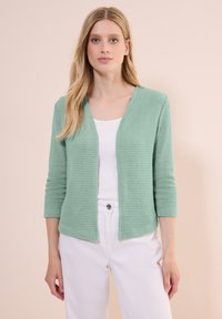 Cardigan texturé vert menthe avec un décolleté en V et des manches trois-quarts, porté sur un haut blanc et assorti avec un pantalon blanc.