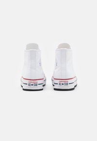 Bílé plátěné vysoké tenisky s černou gumovou podrážkou, červenými a modrými detaily a kruhovým logem na vnější části kotníku.