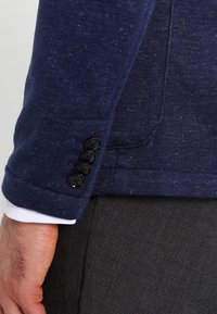 Námořnický modrý blazer s texturovanou tkaninou, se čtyřmi černými knoflíky na manžetě a viditelným bílým rukávem košile pod ním.