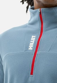 Pull en polaire bleu clair avec une finition texturée, doté d'une fermeture éclair rouge et d'un logo Millet en blanc. Conception à col haut pour plus de chaleur.