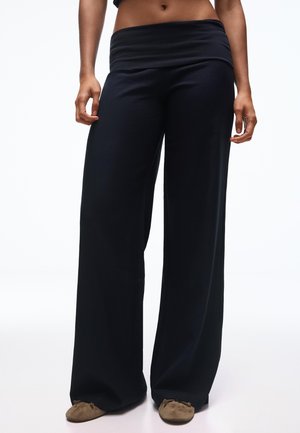 Ženska v širokih, črnih ohlapnih hlačah in rjavih copatih, stoječa pred enobarvnim belim ozadjem.