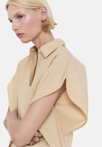 Camisa oversized beige de tela suave con cuello puntiagudo, mangas cortas con un diseño tipo ala y un bolsillo en el pecho como detalle.