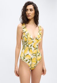 Γυναίκα που φοράει ολόσωμο μαγιό με λαιμόκοψη σε σχήμα V, με κίτρινο σχέδιο λεμονιών και πράσινα φύλλα, βολάν στους ώμους, στέκεται μπροστά σε λευκό φόντο.