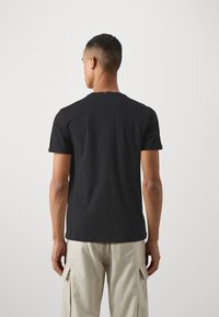 T-shirt en coton noir avec un col rond et des manches courtes, doté d'un petit accent coloré au niveau de l'encolure arrière. Le modèle porte un pantalon cargo beige.