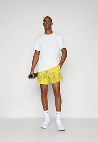 Hombre de pie, vistiendo una camiseta blanca, pantalones cortos amarillos con un patrón, calcetines blancos y zapatillas, sosteniendo una botella de agua negra en la mano derecha.