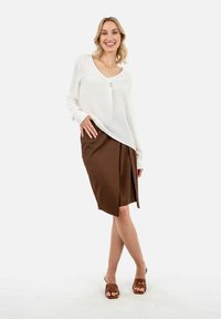 Weiße Bluse mit langen Ärmeln und Knopfdetails, kombiniert mit einem braunen Wickelrock. Braune Riemchensandalen runden das Outfit ab.