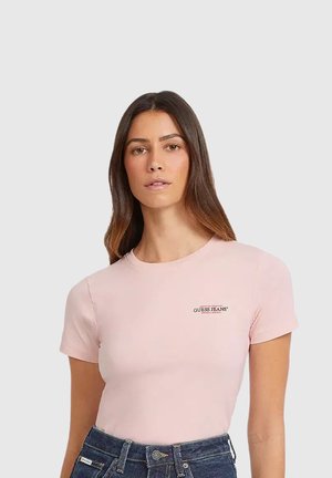 Femme aux longs cheveux bruns portant un t-shirt rose clair Guess Jeans et un jean taille haute bleu foncé, debout devant un fond uni.