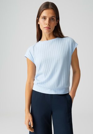 Jeune femme aux longs cheveux bruns portant un haut à manches courtes côtelé bleu clair et un pantalon bleu marine foncé, debout devant un fond uni.