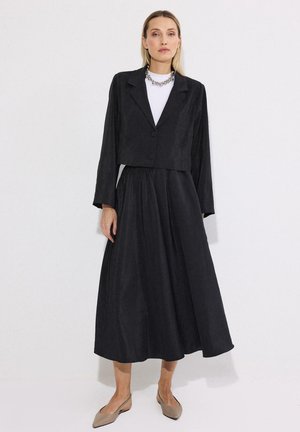 Kobieta w czarnym krótkim blezerze, czarnej spódnicy midi, białym topie, srebrnym naszyjniku i beżowych spiczastych balerinach, stojąca na białym tle.