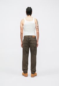 Cargo pants in dark brown fabric with multiple side pockets, worn with a white tank top and brown work boots. Simple design, unembellished.