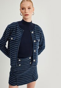 Gestreifte Strickjacke in Marineblau mit silbernen Knöpfen und passendem Rock, mit taillierter Passform und seitlichem Schlitz, kombiniert mit einem dunklen Rollkragenpullover.