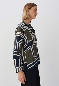 Langsleeve shirt in groen en marineblauw met geometrische witte patronen. Gladde textuur, kraagdetails en zijsplitten aan de onderrand.