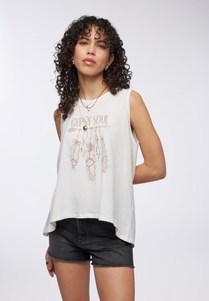Top bianco senza maniche con la scritta "Gypsy Soul" e una grafica di una piuma marrone. Abbinato a pantaloni corti di jeans neri con orlo sfilacciato.