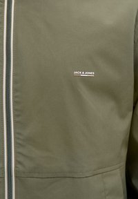 Kurtka w oliwkowej zieleni z przednim zamkiem błyskawicznym i małym białym logo "JACK & JONES" na piersi.